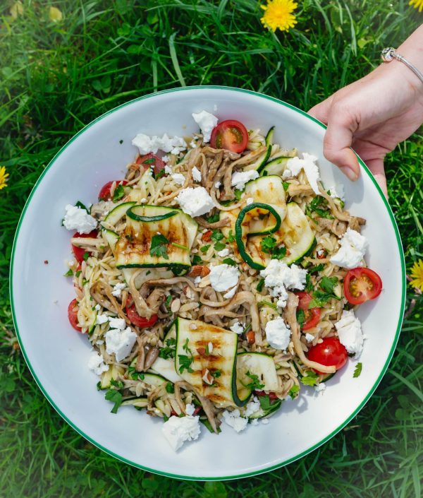 Ingrediënten voor orzo salade: oesterzwammen, courgette, cherrytomaatjes, feta, peterselie, citroen en kruiden op een aanrecht